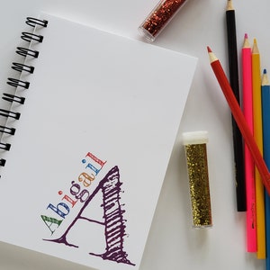 Personalized notebook with white cover and a colorful name and monogram initial in the lower right side of the cover. These custom notebooks are great for girls or boys, and women. Perfect gift idea for party favors, students and teachers.