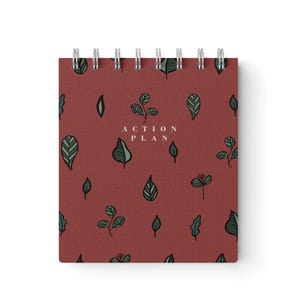 May include: A spiral-bound notebook with a burgundy cover featuring a pattern of green leaves. The words "ACTION PLAN" are printed in white. The notebook is designed for planning and organization.