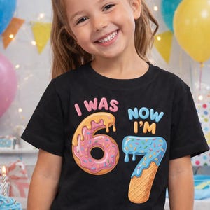 Puede incluir: Una camiseta negra con el texto "I WAS 6, NOW I'M 7" en letras coloridas. El "6" está diseñado como una rosquilla y el "7" como un cono de helado. La camiseta la lleva un niño sonriente.