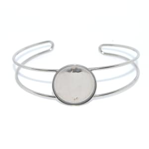 Puede incluir: Un brazalete de plata con un engaste redondo, de color plateado, en blanco. El engaste tiene un diámetro de aproximadamente 25 mm.