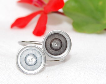 May include: A silver-toned ring with two circular settings. The settings have a metallic appearance with a central recessed area. The ring is displayed against a white background, with a red flower and green leaf in the background.
