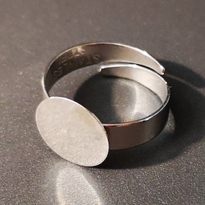 Może przedstawiać: Srebrny regulowany pierścień z okrągłą, pustą, srebrną oprawą. Pierścień jest oznaczony "STAINLESS STEEL" od wewnątrz.