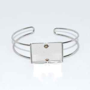 Puede incluir: Un brazalete de plata con un panel de vidrio cuadrado en el centro. El panel de vidrio es transparente y tiene un marco plateado.