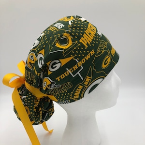 May include: Green and gold surgical scrub hat with a Green Bay Packers theme. The hat has a tie on the back and features the team logo, the words "Packers", "Touchdown", and "Green Bay".