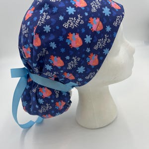 May include: Blue surgical scrub hat with a white and pink heart pattern and the text "Bless your Heart" repeated throughout. The hat has a blue ribbon tie.