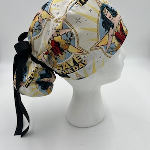 May include: White surgical scrub hat with a Wonder Woman print. The hat features a repeated pattern of Wonder Woman in a star with the text "Save the Day" and "The Day". The hat has a black tie.