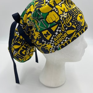 May include: A yellow, green, and blue patterned surgical scrub hat with a black ribbon tie. The hat features the text "Hail", "Michigan", and "Ann Arbor".
