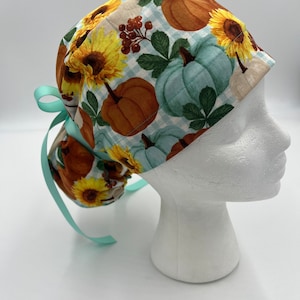 May include: A turquoise and white patterned surgical scrub hat with a tie. The pattern features pumpkins, sunflowers, and leaves.