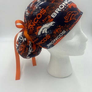 Op de afbeelding: Een blauwe en oranje chirurgische scrub-muts met een strik aan de achterkant. De muts heeft een herhalend patroon van het Denver Broncos-logo en de woorden "Broncos" en "Denver".