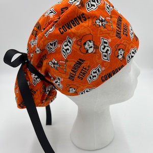 Peut inclure: Un bonnet de travail orange et noir avec un motif du logo de l'Université d'État de l'Oklahoma et les mots "Cowboys" et "Oklahoma State". Le bonnet a des liens noirs.