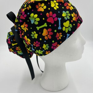 Op de afbeelding: Zwarte scrub-muts met een kleurrijk patroon van pootjes en botjes.