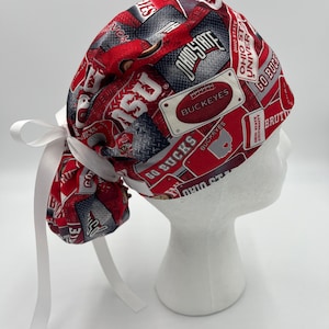 以下が含まれることがあります： 白の紐が付いた赤とグレーの柄の外科用スクラブキャップ。パターンには「オハイオ州立大学」、「バッカイズ」、「ゴーバックス」の文字が書かれています。