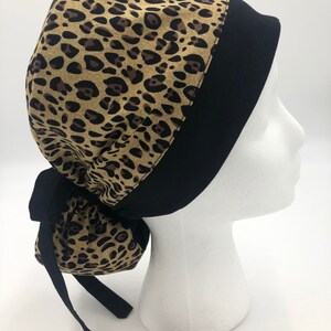 May include: A leopard print surgical scrub hat with a black trim and tie. The hat is tied in a knot at the back of the head.
