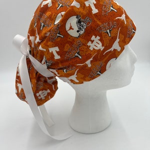 May include: Orange and white scrub hat with a Texas Longhorns pattern. The hat has a white tie that can be tied at the back of the head.