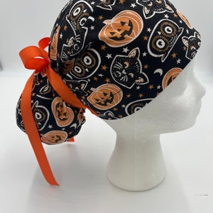 Puede incluir: Un gorro de quirófano negro con un estampado de Halloween naranja y negro con calabazas, gatos y estrellas. El gorro tiene una cinta naranja larga atada en la parte posterior.