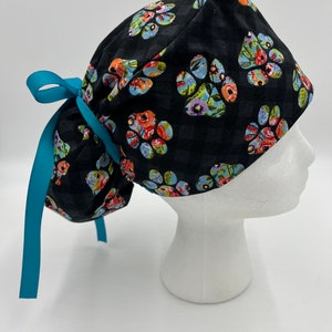 Op de afbeelding: Een zwarte scrub-muts met een kleurrijk pootprintpatroon en een turquoise lint.