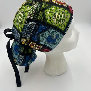 May include: A patterned scrub cap featuring a stained glass design with green, blue, and red accents. The cap includes the text "SLYTHERIN" and a black tie in the back. The cap is displayed on a white mannequin head.