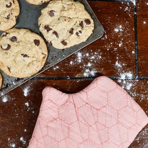 May include: A metal baking tray holding several round, golden-brown chocolate chip cookies, positioned next to a light pink oven glove with a textured hexagonal pattern. White flour is scattered across the dark brown wooden surface.