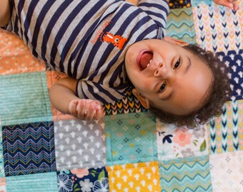 May include: A baby wearing a striped blue and gray onesie with a small fox graphic on the front, lying on a colorful patchwork quilt.