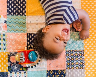 May include: A baby lying on a colorful patchwork quilt, laughing and looking up at the camera. The baby is wearing a striped shirt and has curly brown hair. A colorful toy with a black and white handle is in the foreground.