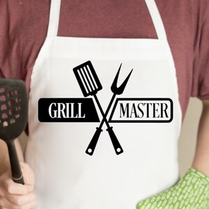 May include: A white apron featuring a black graphic print of a spatula and a two-pronged fork crossed behind the text "GRILL MASTER" in a bold, sans-serif font. The apron has a neck loop and waist ties, providing coverage for the torso area.