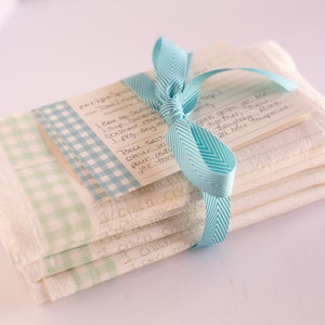 May include: A stack of white kitchen towels with a blue and white checkered pattern are tied together with a blue ribbon. The top towel has a handwritten recipe card attached to it.