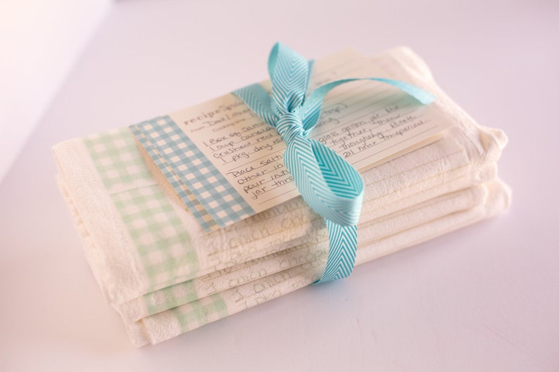 May include: A stack of white kitchen towels with a blue and white checkered pattern are tied together with a blue ribbon. The top towel has a handwritten recipe card attached to it.