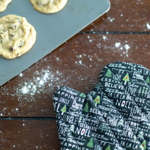 Könnte beinhalten: Ein schwarzer und grüner Ofenhandschuh mit gesteppter Textur und weißem Text, der "Let it Snow", "Peace on Earth", "Believe", "Love", "Noel", "Merry Christmas", "Fa La La" und "Jingle" auf einer braunen Holzoberfläche mit weißem Pulver sagt.