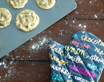 May include: A baking sheet with freshly baked chocolate chip cookies on a wooden surface. A gray oven mitt with colorful text is in the foreground. The text on the oven mitt reads "chocolate chip, lemon, vanilla, mango, strawberry, mint, blueberry, coconut".