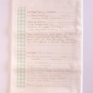 May include: A handwritten recipe for Spicy Crackers and Taco Soup on a white kitchen towel with a green and white checkered border. The recipe for Spicy Crackers includes ingredients like Saltines, canola oil, crushed red pepper, and dry ramen dressing. The recipe for Taco Soup includes ingredients like hamburger, chili, taco mix, ranch style dressing mix, corn, stewed tomatoes, and ranch style beans.