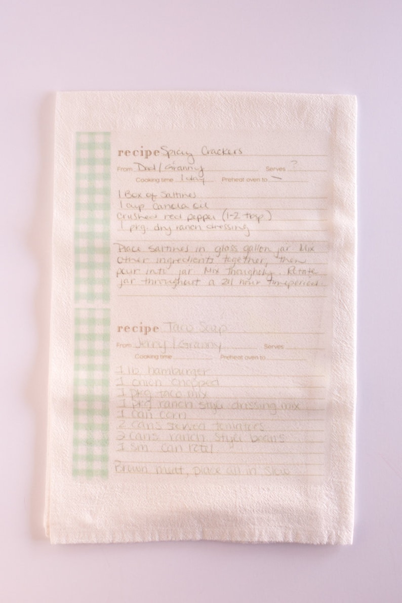 May include: A handwritten recipe for Spicy Crackers and Taco Soup on a white kitchen towel with a green and white checkered border. The recipe for Spicy Crackers includes ingredients like Saltines, canola oil, crushed red pepper, and dry ramen dressing. The recipe for Taco Soup includes ingredients like hamburger, chili, taco mix, ranch style dressing mix, corn, stewed tomatoes, and ranch style beans.