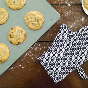 Könnte beinhalten: Ein Backblech mit frisch gebackenen Schokoladen-Chip-Cookies auf einer Holzoberfläche. Ein schwarz-weißes Ofenhandschuh mit sechseckigem Muster befindet sich ebenfalls auf der Oberfläche.