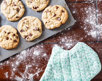 May include: A baking sheet holds freshly baked chocolate chip cookies. The cookies are golden brown with visible chocolate chips. Flour is dusted on the dark wooden surface, and a green floral oven mitt is in the corner.