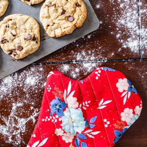 Puede incluir: Un guante de horno rojo con un patrón floral, sentado en una mesa de madera con una bandeja para hornear galletas con chispas de chocolate y harina esparcida alrededor.