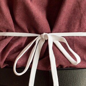 May include: A white fabric apron tie fastened in a bow at the waist over a maroon t-shirt and a black belt. The apron strings are thin and flat, creating gathered folds in the fabric of the shirt beneath the tie, centered against the torso.