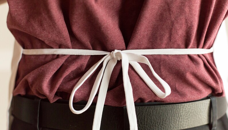 May include: A white fabric apron tie fastened in a bow at the waist over a maroon t-shirt and a black belt. The apron strings are thin and flat, creating gathered folds in the fabric of the shirt beneath the tie, centered against the torso.