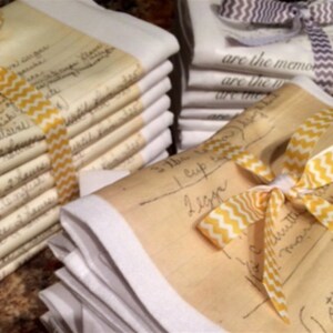 May include: A stack of white tea towels with handwritten recipes printed on them. The towels are tied together with yellow and white ribbon.