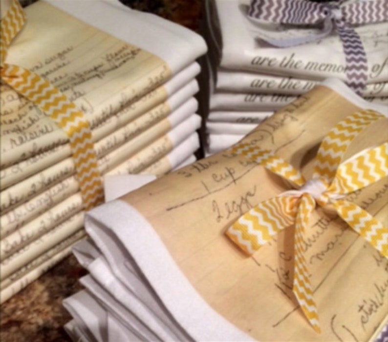 May include: A stack of white tea towels with handwritten recipes printed on them. The towels are tied together with yellow and white ribbon.