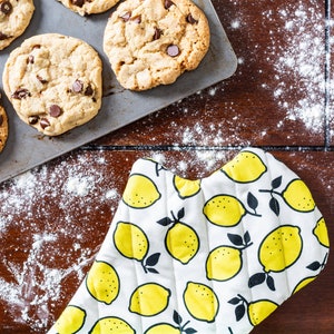 May include: A baking sheet with chocolate chip cookies on it. The cookies are on a brown wooden surface with a white oven mitt featuring a lemon pattern.