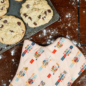 Puede incluir: Toma cenital de galletas con chispas de chocolate recién horneadas en una bandeja de metal para hornear junto a un guante de horno rosa con un patrón repetido de cascanueces. Harina espolvoreada sobre la superficie de madera oscura.