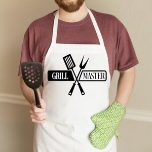 May include: A white apron featuring a black graphic of a crossed spatula and carving fork with the text "GRILL MASTER" printed in the center. The apron includes a neck strap and waist ties, providing full coverage for the front of the torso.