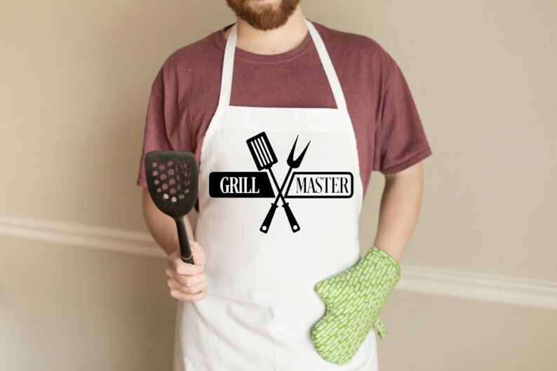 May include: A white apron featuring a black graphic of a crossed spatula and carving fork with the text "GRILL MASTER" printed in the center. The apron includes a neck strap and waist ties, providing full coverage for the front of the torso.