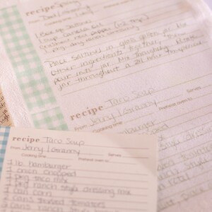 May include: Two recipe cards with a blue and white checkered pattern on the front. The recipe cards are for Taco Soup. The first card is from Jerry I. Grammy and includes ingredients like hamburger, onion, corn, and ranch style dressing mix. The second card is from "Granny" and includes ingredients like salsa, crushed red pepper, and dry ranch.