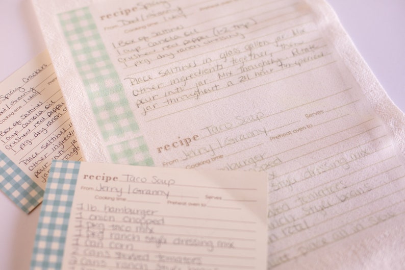 May include: Two recipe cards with a blue and white checkered pattern on the front. The recipe cards are for Taco Soup. The first card is from Jerry I. Grammy and includes ingredients like hamburger, onion, corn, and ranch style dressing mix. The second card is from "Granny" and includes ingredients like salsa, crushed red pepper, and dry ranch.