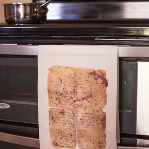 May include: A white kitchen towel hanging on a stainless steel oven door. The towel has a vintage, aged piece of paper with handwritten text and brown and red stains hanging from it.