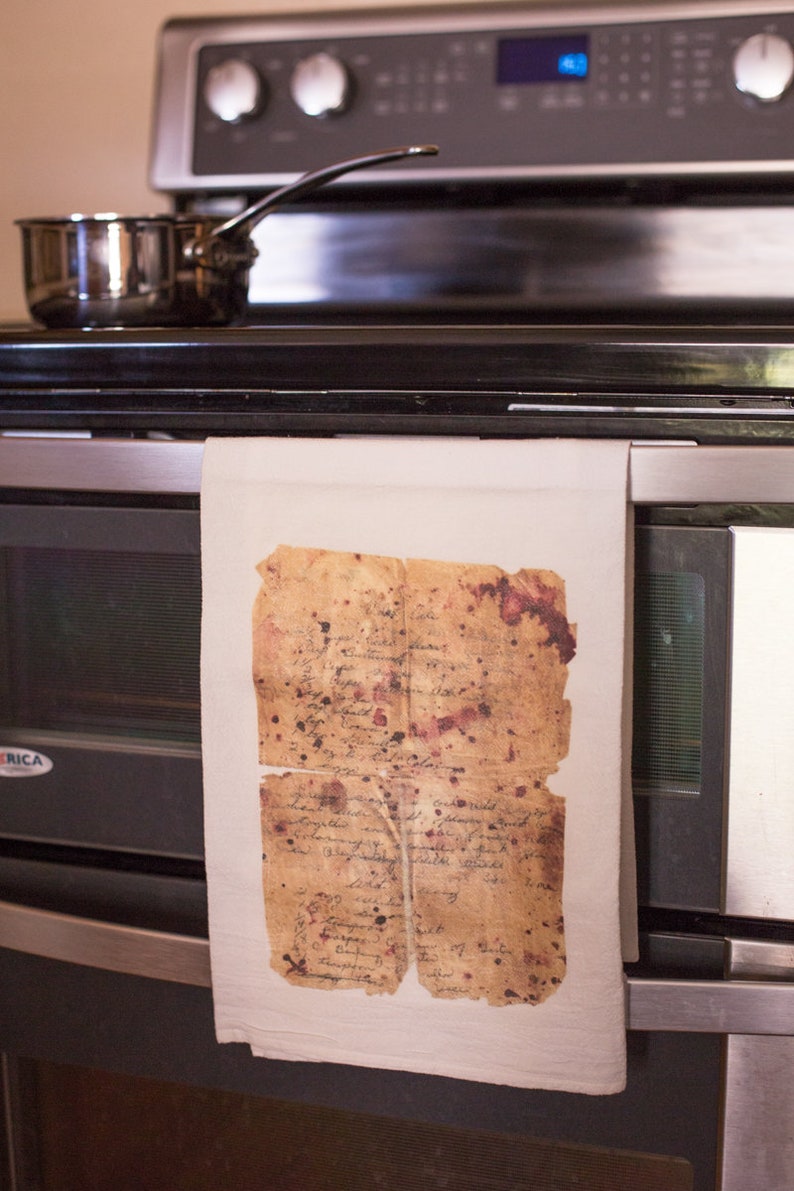 May include: A white kitchen towel hanging on a stainless steel oven door. The towel has a vintage, aged piece of paper with handwritten text and brown and red stains hanging from it.