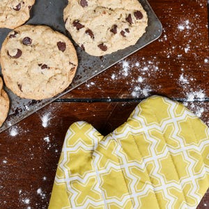May include: Overhead shot of freshly baked chocolate chip cookies on a baking sheet, alongside a yellow oven mitt with a white geometric pattern. Flour is sprinkled on the dark wooden surface.