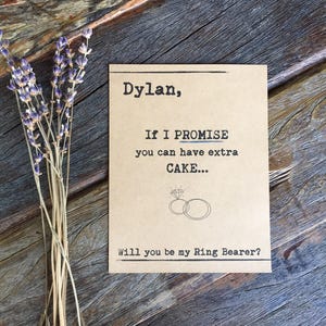 May include: A brown card with the text "Dylan, If I PROMISE you can have extra CAKE... Will you be my Ring Bearer?" with a drawing of two rings with a diamond on top.