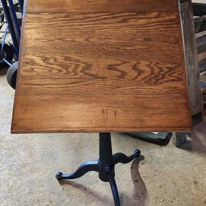May include: A wooden lectern with a slanted top featuring a prominent brown wood grain pattern. The lectern is supported by a central black pedestal base that branches out into three curved legs at the bottom, providing a stable foundation.