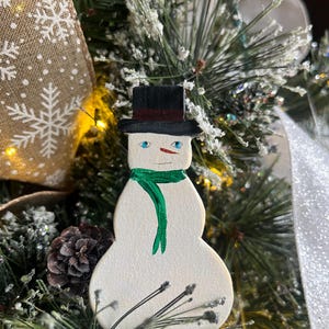 May include: A white snowman ornament with a black top hat, red trim, green scarf, and blue eyes. The ornament is hanging on a Christmas tree.